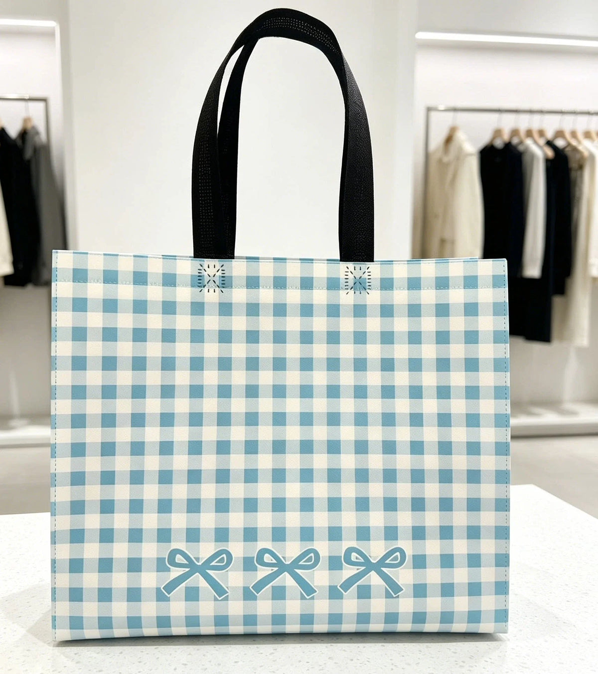 Blue gingham laminated non-woven tote with black loop handles and bow artwork in a clothing store